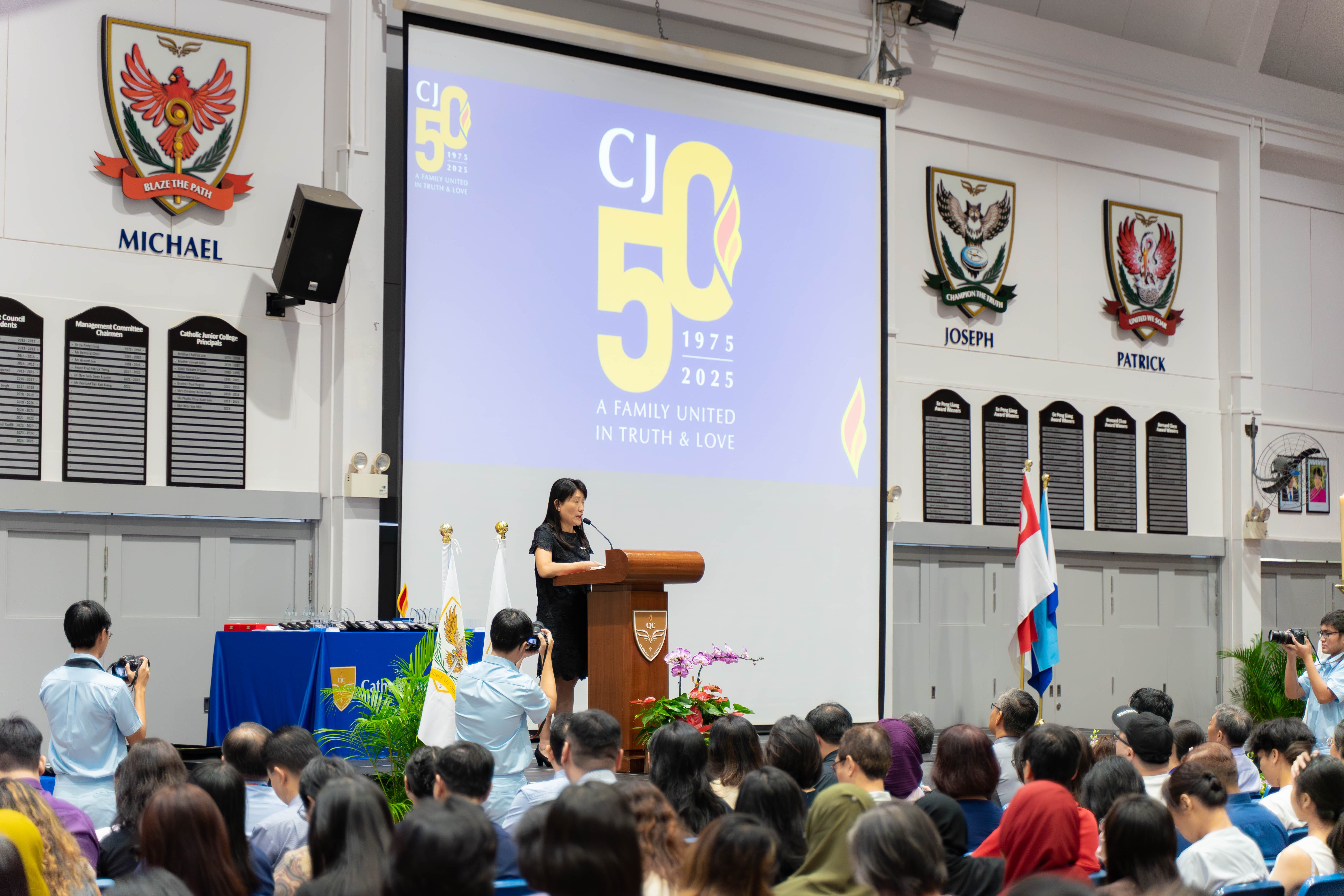A woman speaks at a podium during Catholic Junior College's 50th anniversary event, with a large CJ50 screen behind her.